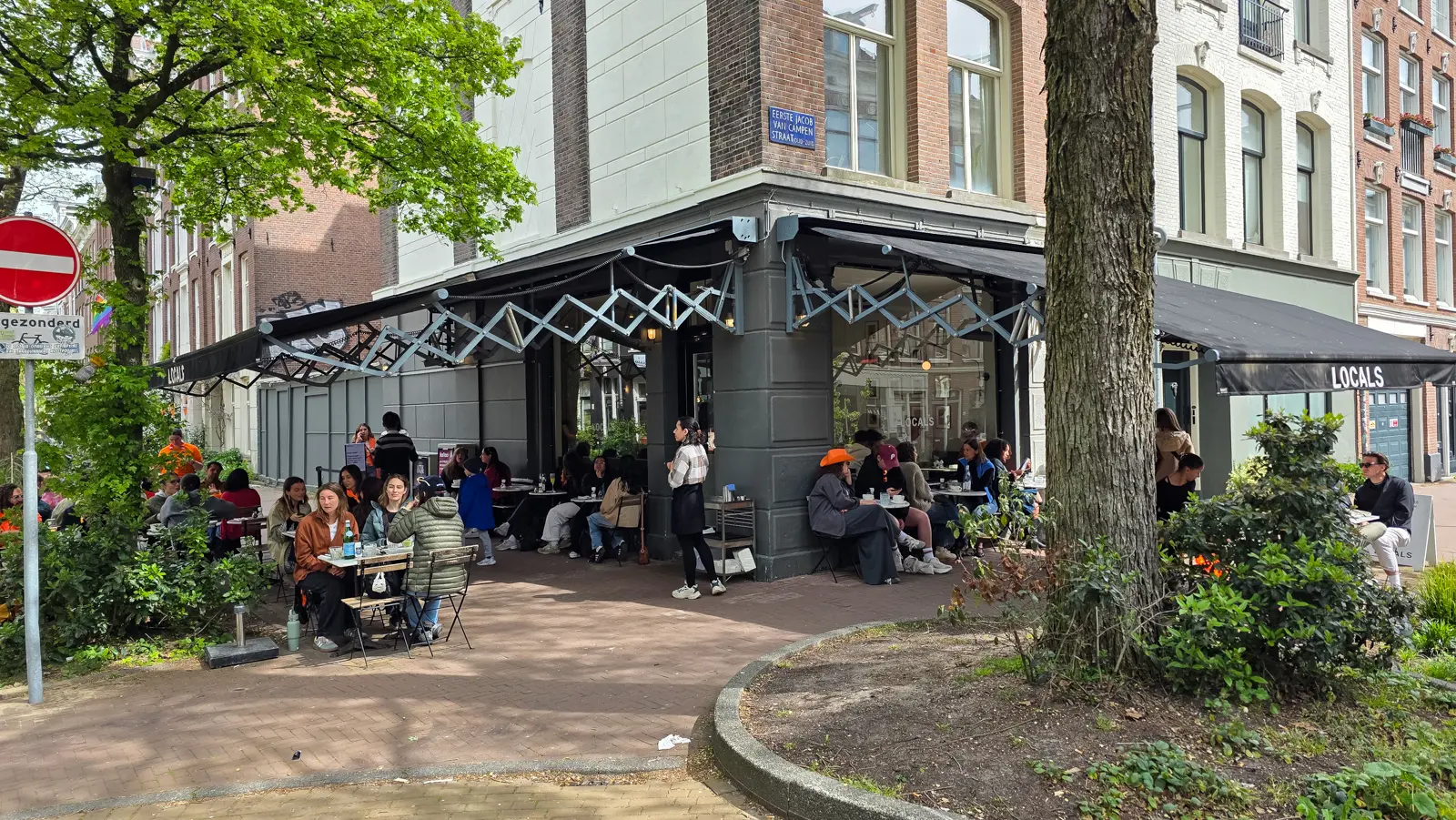 Corner exterior of Locals in De Pijp, Amsterdam, with black awnings, pavement seating and the blue street sign above