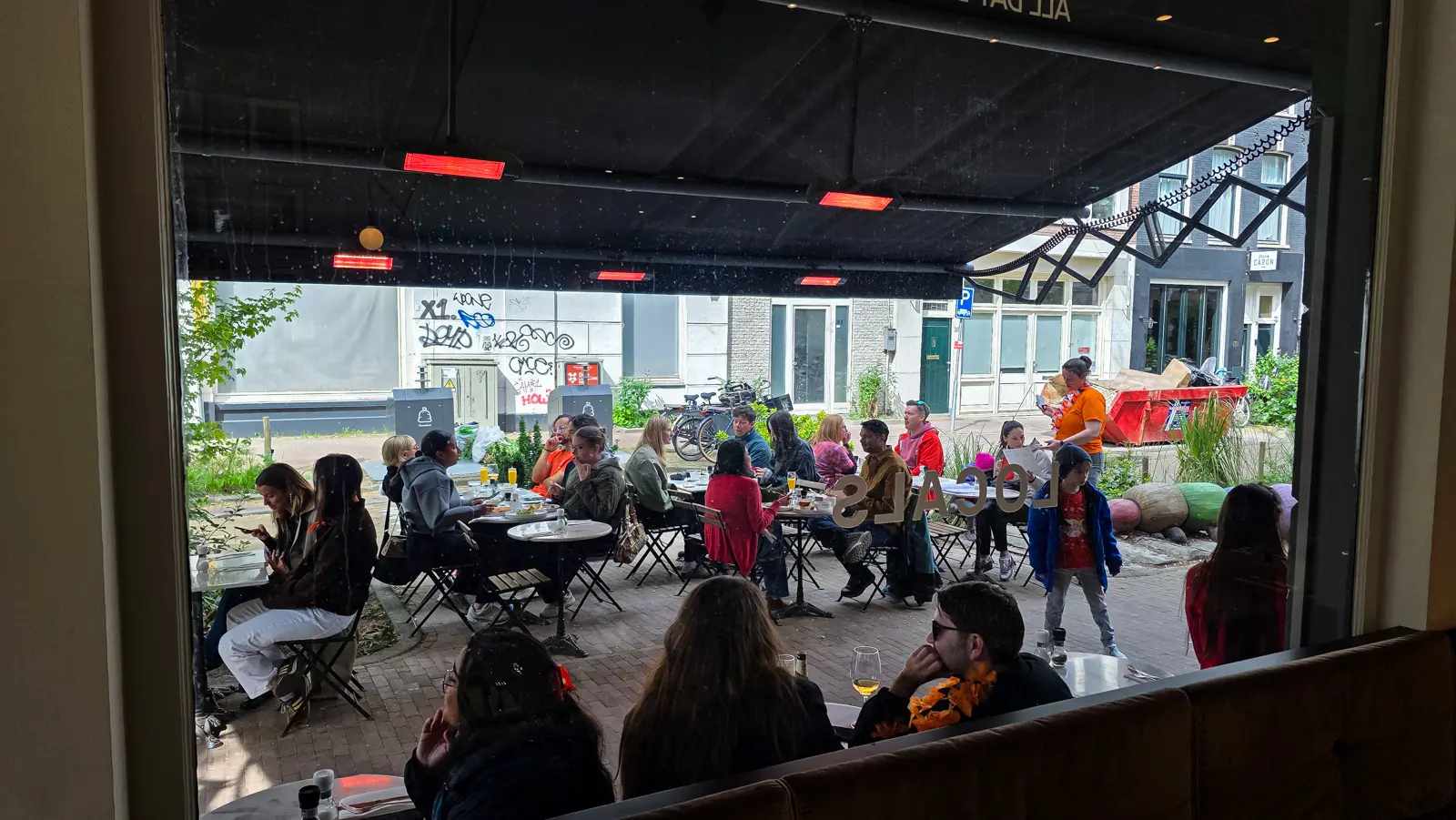 The covered terrace at Locals Amsterdam seen from inside, tables full, infrared heaters glowing under the black awning