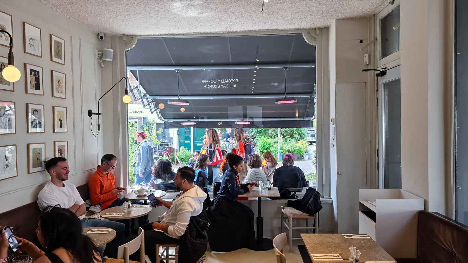 Interior of Locals in De Pijp, Amsterdam, with framed drawings covering one wall, stone tables and an open frontage onto the street