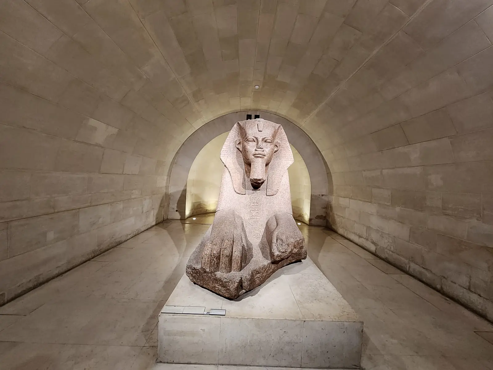 The Great Sphinx of Tanis in a vaulted stone crypt at the Louvre, lit from below, its paws forward and headdress framed by the arch
