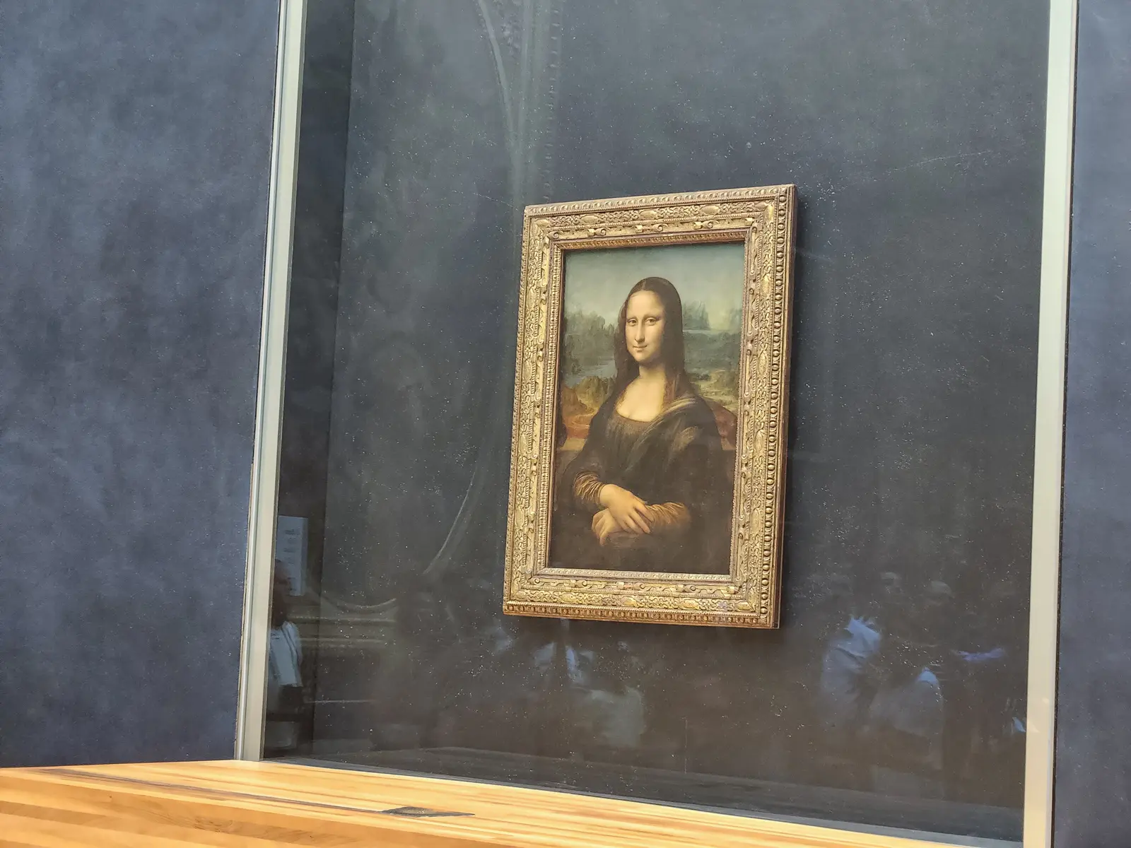 The Mona Lisa in its gilded frame behind glass on a dark wall at the Louvre, with reflections of visitors across the surface
