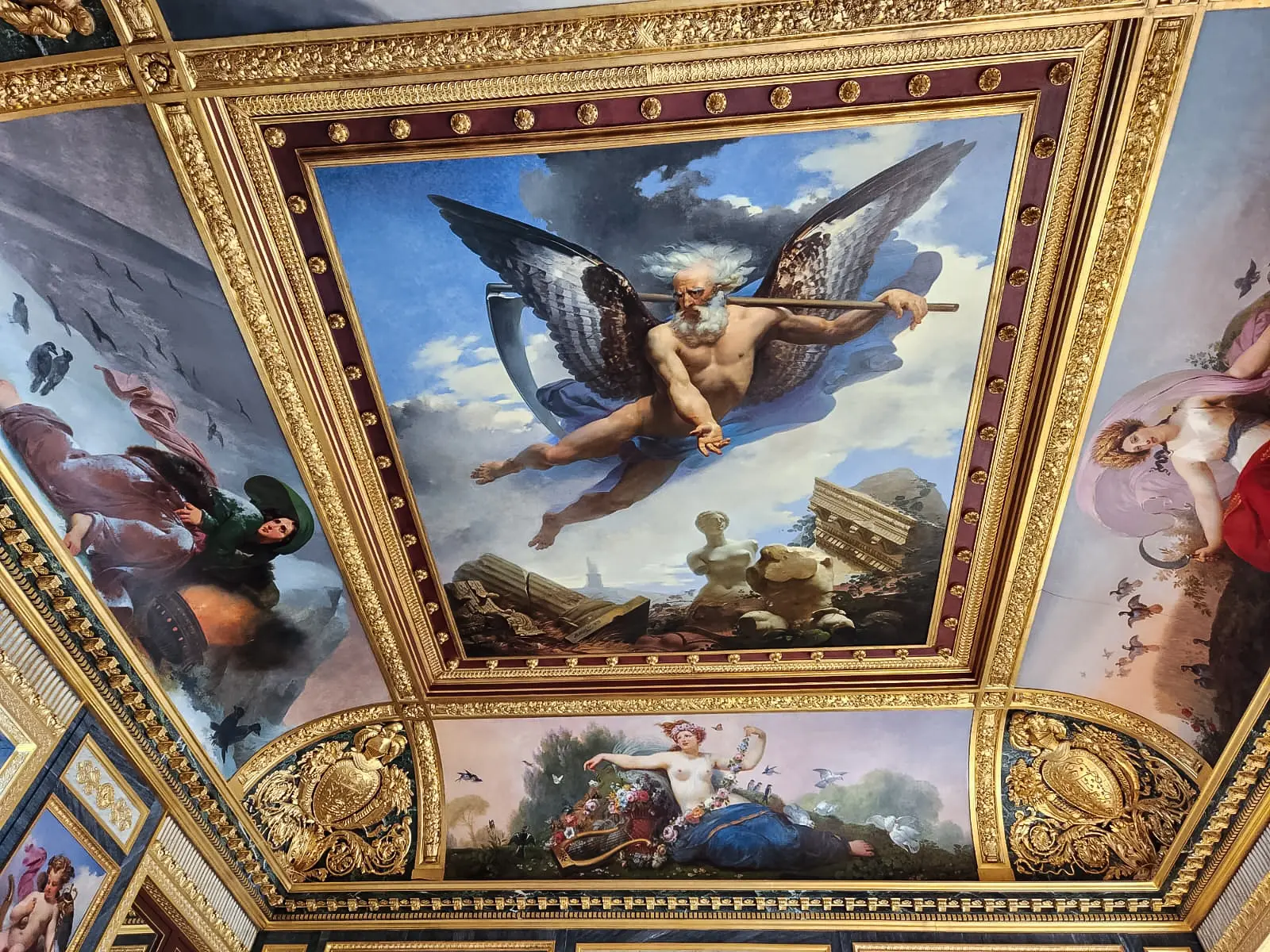 A painted and gilded Louvre ceiling showing a winged bearded figure with a scythe flying above classical ruins in a heavy frame