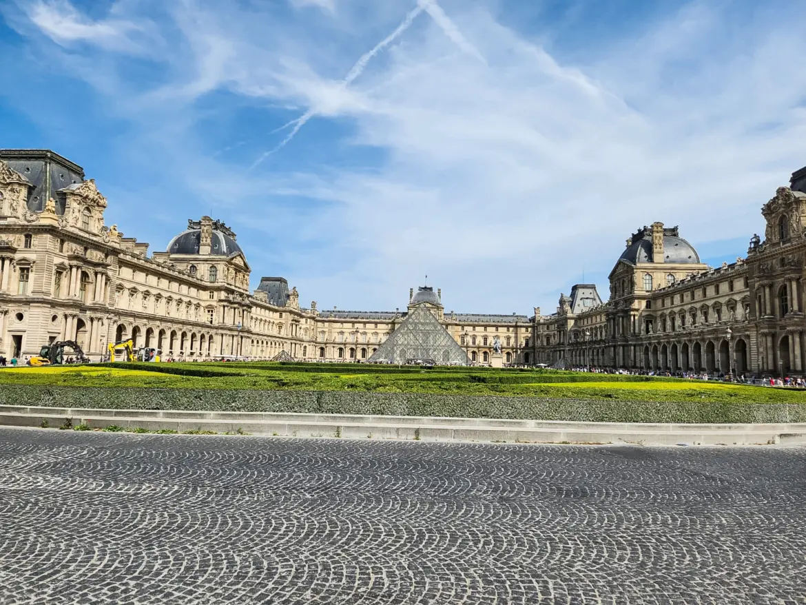 The Louvre’s courtyard wings framing the glass pyramid, seen across cobbles and a low hedge under a wide blue sky in Paris