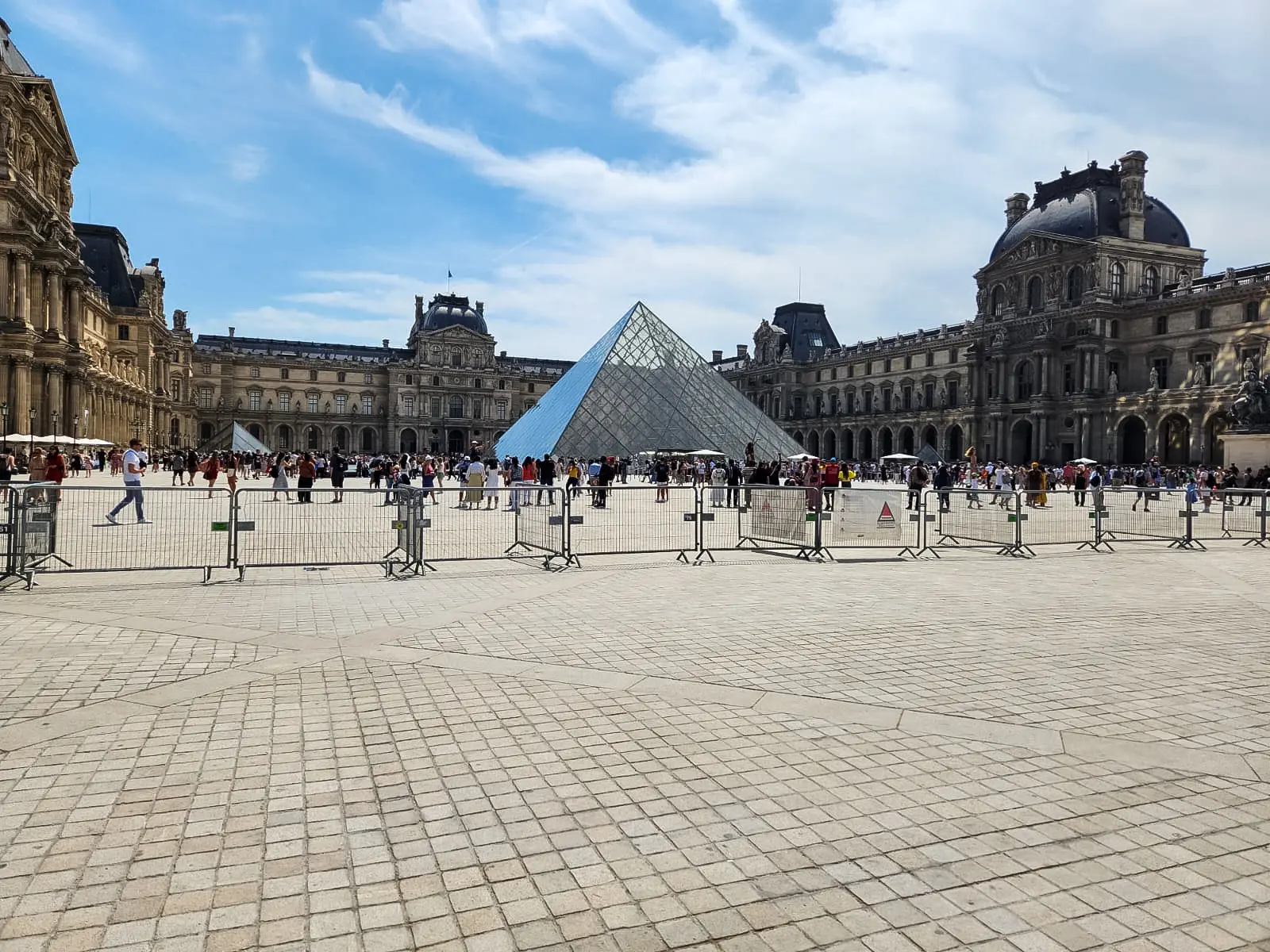 Crowds gathered around the glass pyramid in the Louvre courtyard behind metal barriers, with the palace wings on either side