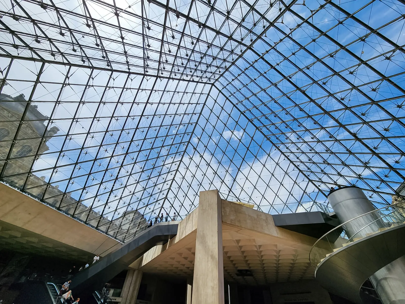 Looking up through the glass and steel lattice of the Louvre pyramid from inside the entrance hall, with sky and palace visible beyond