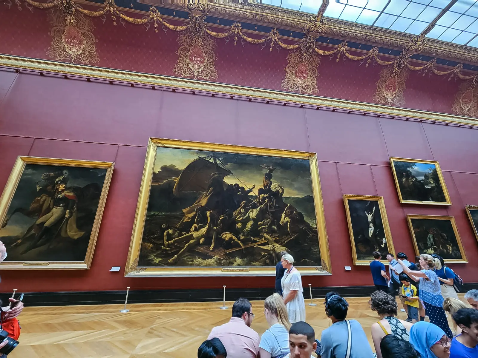 Géricault’s Raft of the Medusa hanging on a deep red wall at the Louvre among gilded frames, with visitors gathered on the parquet below