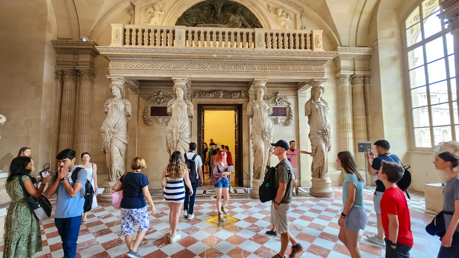 Visitors on the red and white marble floor of the Salle des Caryatides at the Louvre, below four carved figures holding up a gallery