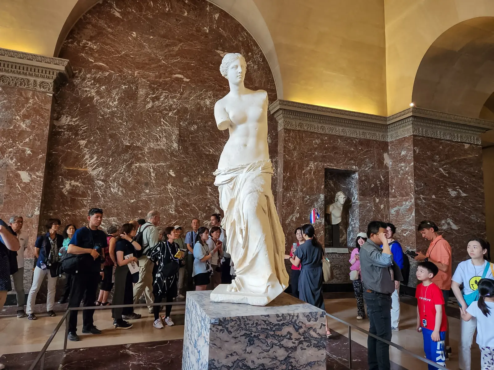 The Venus de Milo on a marble plinth in a red-walled hall at the Louvre, encircled by visitors taking photographs
