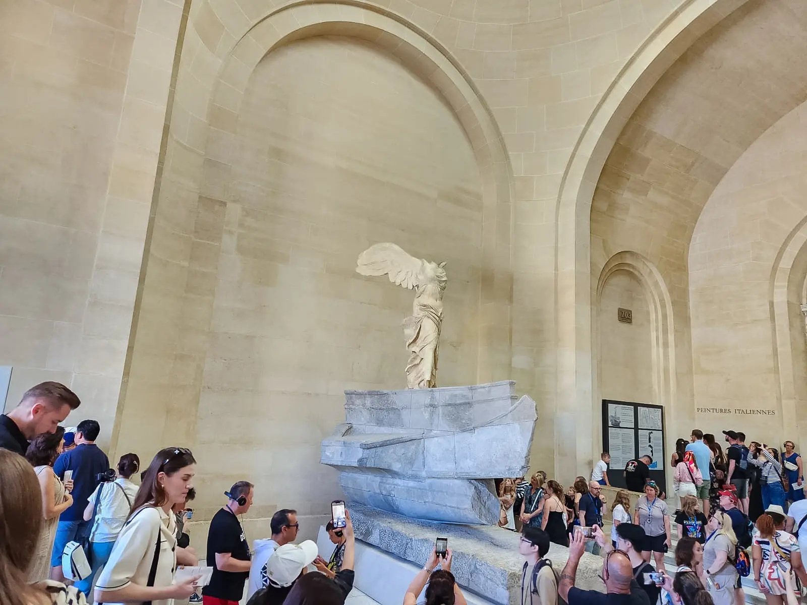 The Winged Victory of Samothrace seen above a dense crowd of visitors holding phones on the staircase below her at the Louvre