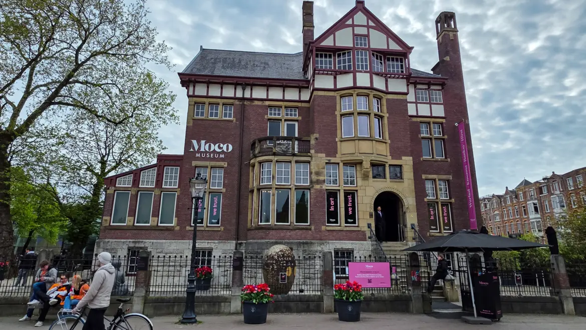 The Moco Museum in Amsterdam, a brick and half-timbered villa on Museumplein with pink “In art we trust” banners, under a grey sky