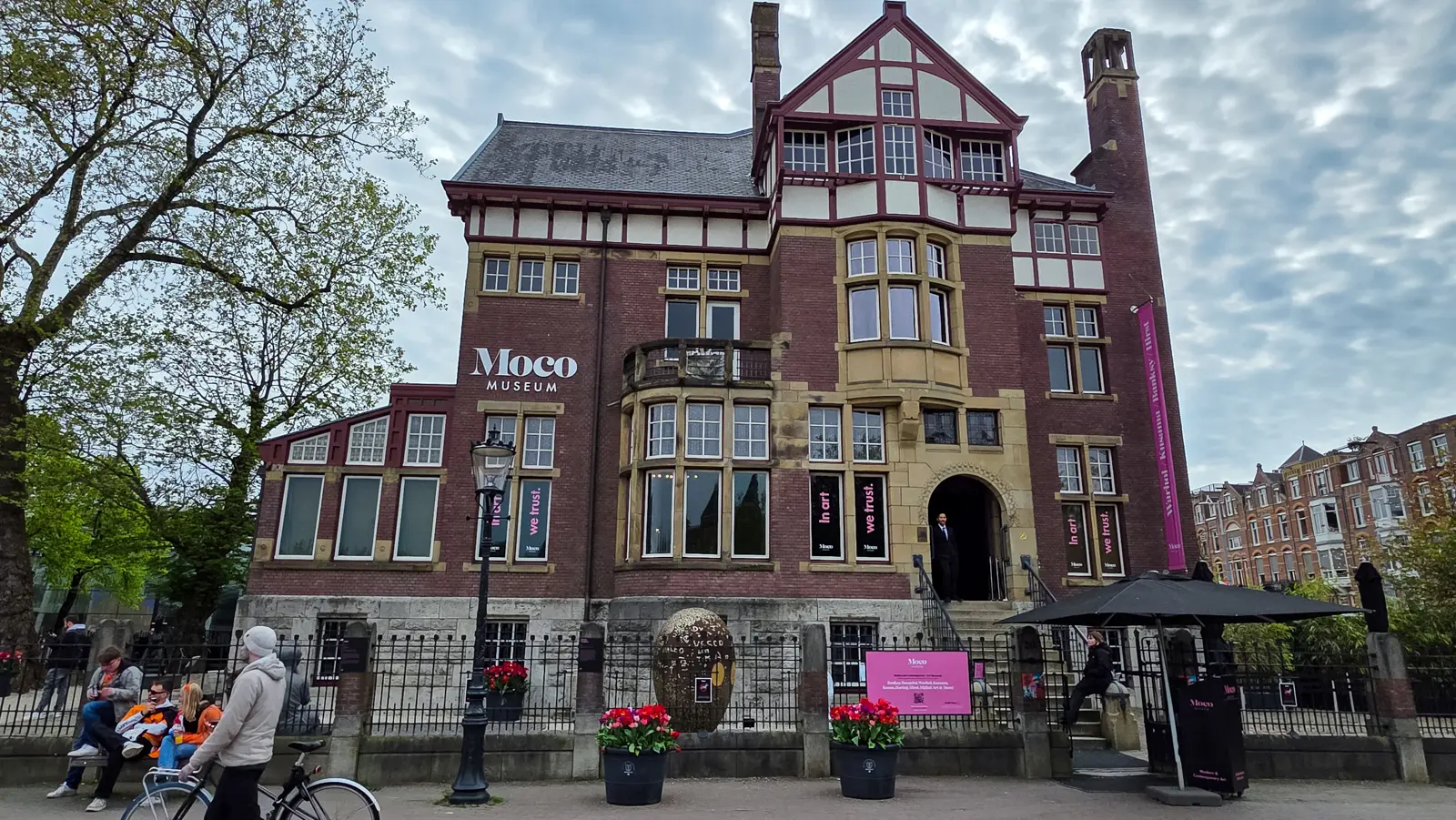 The Moco Museum in Amsterdam, a brick and timbered villa on Museumplein with pink In art we trust banners, under a grey sky