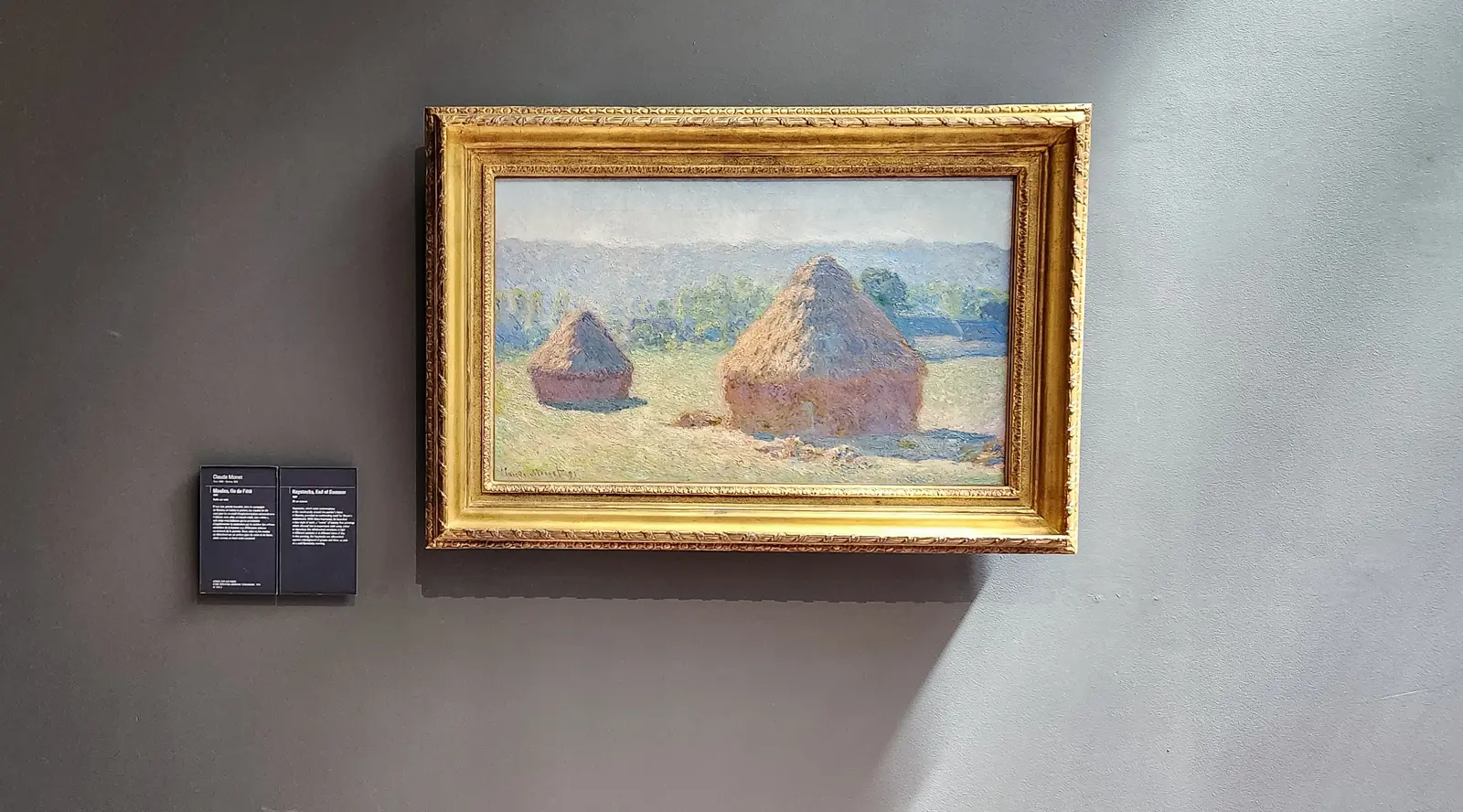 Monet’s Haystacks, End of Summer at the Musée d’Orsay, two stacks in a pale field under a soft sky, in a gilt frame on a grey wall