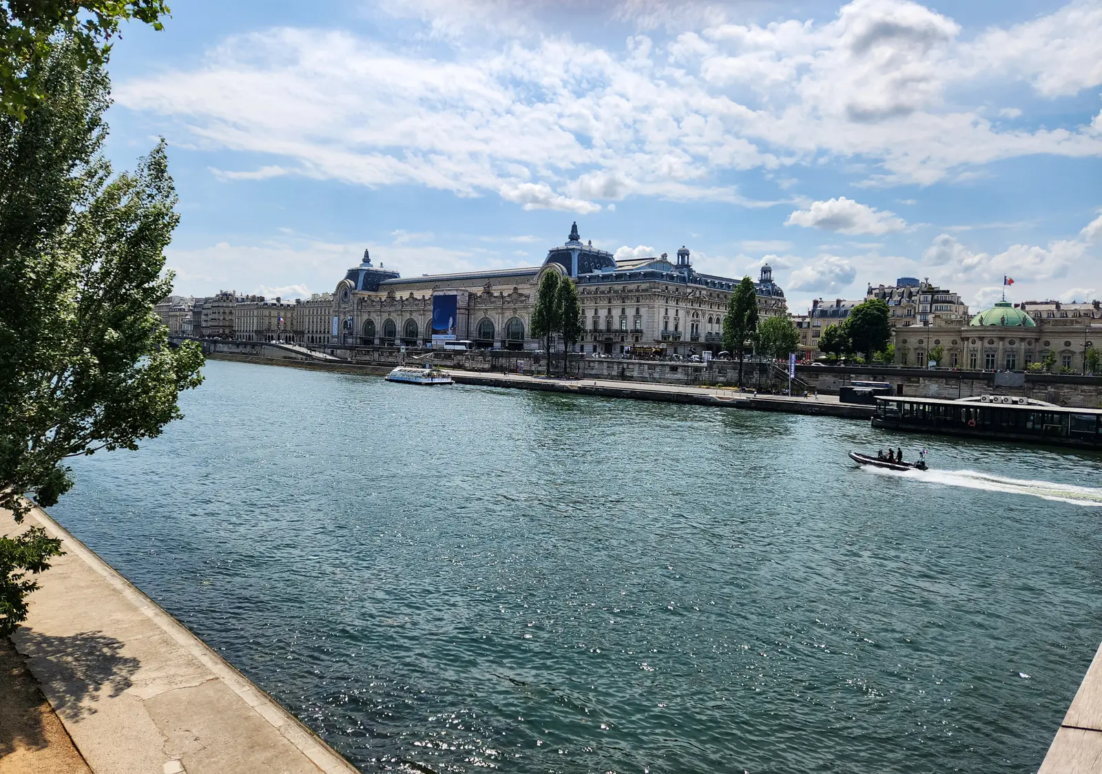 The Musée d’Orsay seen across the Seine on a bright day, its long stone facade and clock faces above the quay, with boats on the water