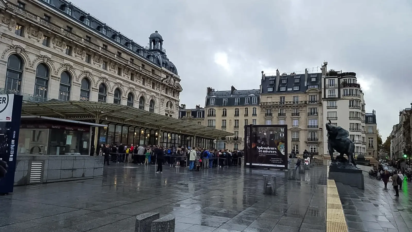 The Musée d’Orsay from the square on a wet grey day, with a queue under the entrance canopy and a bronze animal sculpture at the right