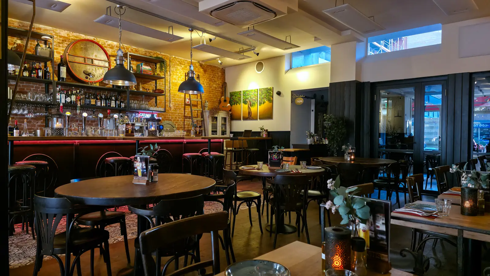 The bar at Oresti’s Taverna in Amsterdam, exposed brick with a mounted wine barrel, hanging lamps, a guitar and a triptych of a tree behind the counter