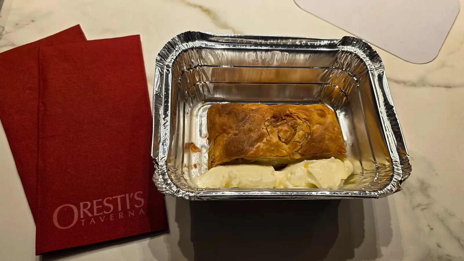 A takeaway bougatsa in a foil tray with cream beside it, resting on a red napkin printed Oresti’s Taverna, on a marble surface