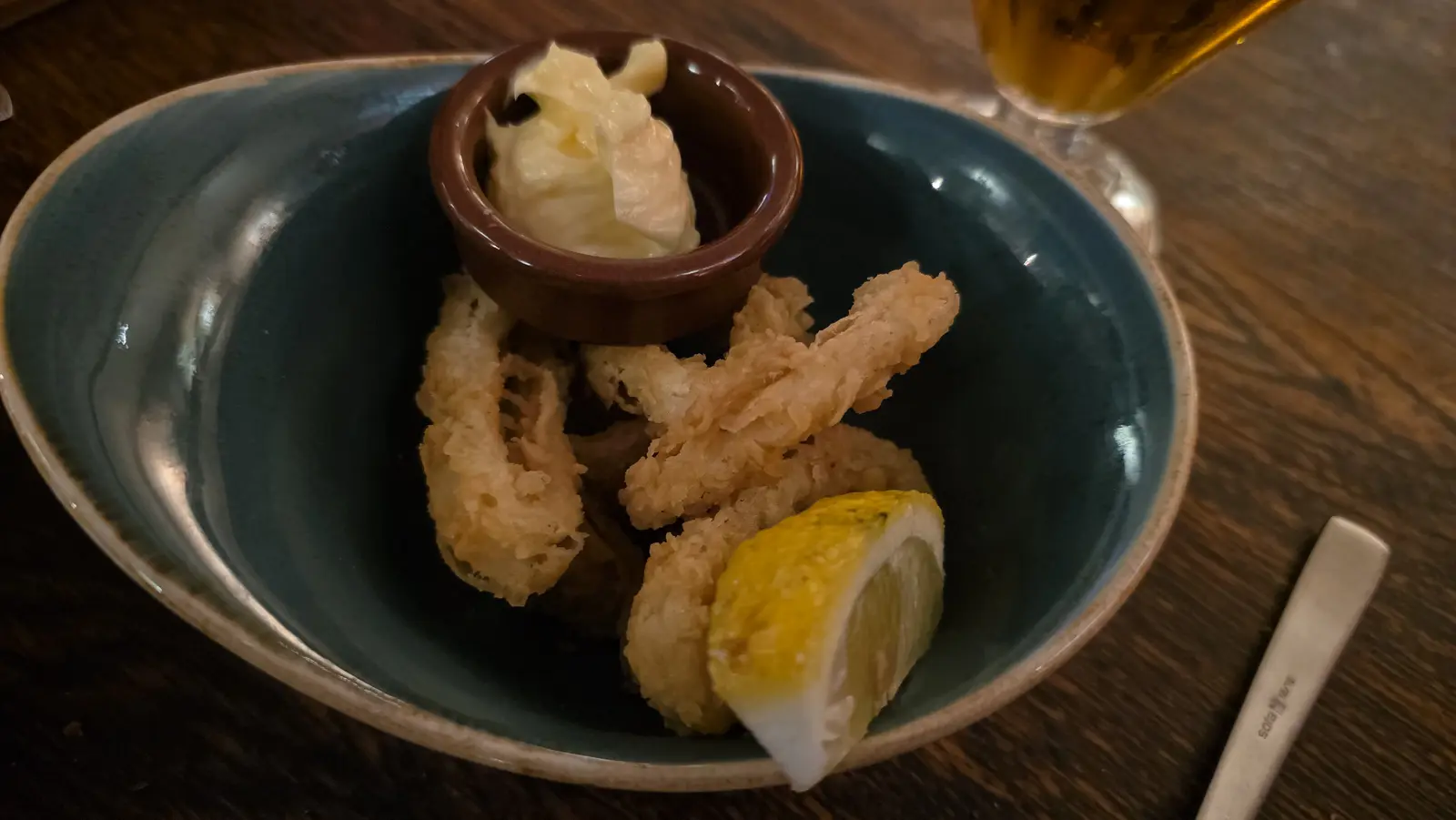 Deep-fried squid rings with a pot of garlic sauce and a lemon wedge in a dark bowl at Oresti’s Taverna
