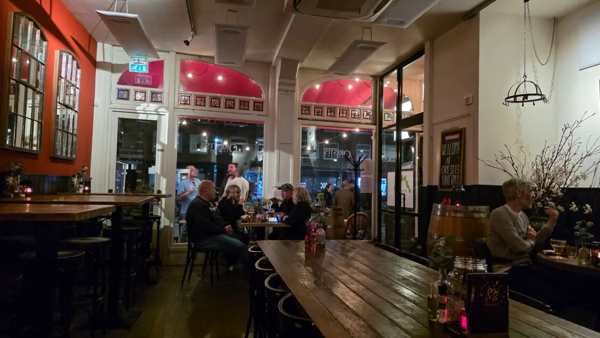 The dining room at Oresti’s Taverna in Amsterdam at night, a welcome board on the wall, stained glass, a wine barrel and diners at wooden tables