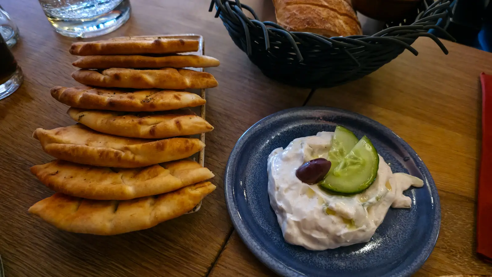 A stack of warm pita breads beside a bowl of tzatziki topped with cucumber slices and an olive, at Oresti’s Taverna