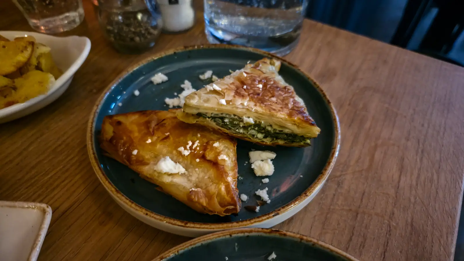 Two triangles of spanakopita on a plate at Oresti’s Taverna, one cut open to show the spinach and feta filling, with feta scattered on the plate