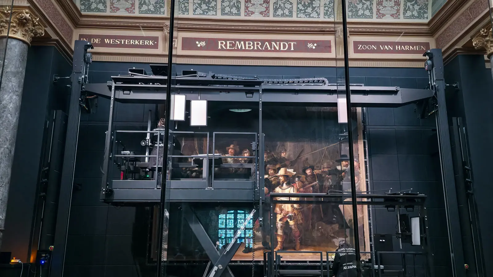 Rembrandt’s Night Watch inside its glass restoration chamber at the Rijksmuseum, with a steel gantry, lighting panels and conservators