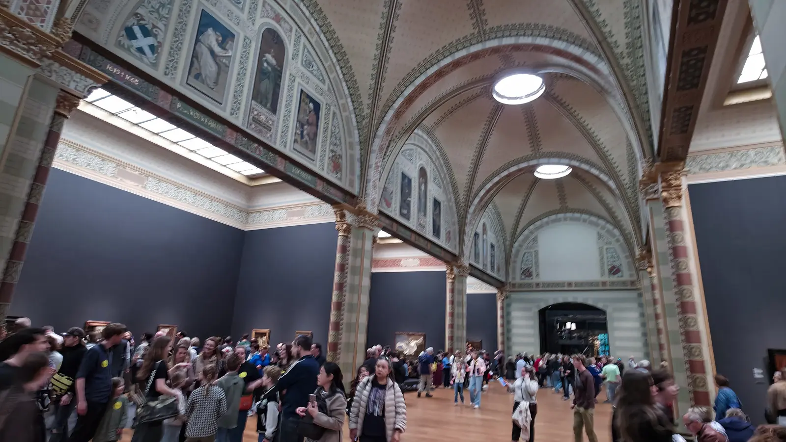 The Gallery of Honour at the Rijksmuseum, with painted vaulted ceilings and skylights above a crowd of visitors moving down the arcade