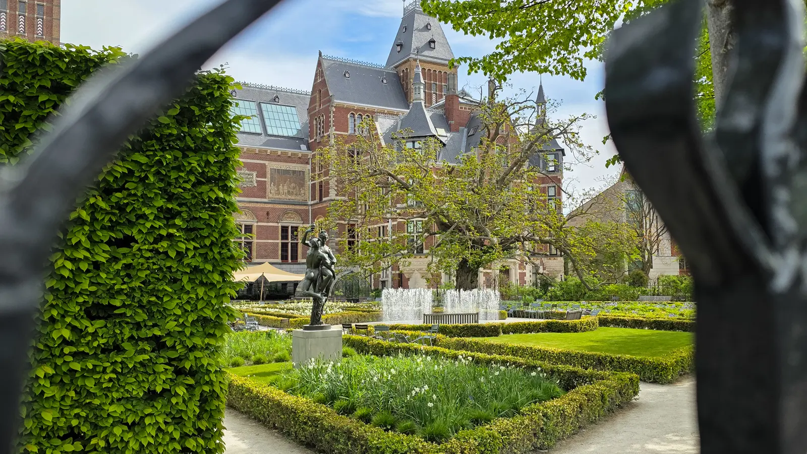 The Rijksmuseum gardens seen through iron railings, with clipped hedges, a fountain, a bronze figure on a plinth and the museum behind