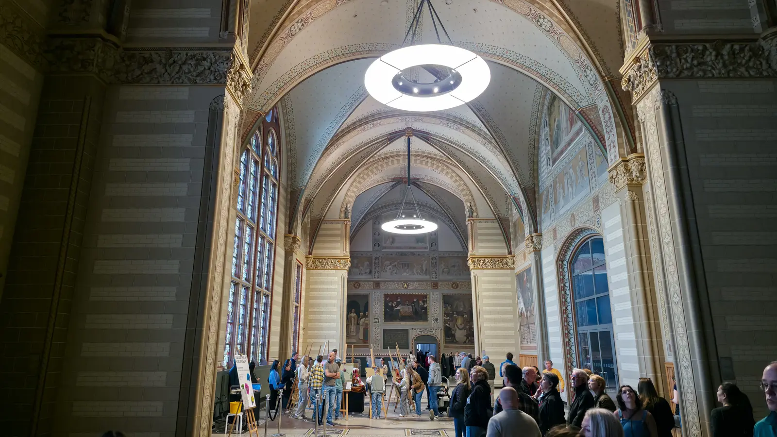 The Great Hall at the Rijksmuseum in painted stone and stencilled ornament, with ring lights above and easels set up on the floor below
