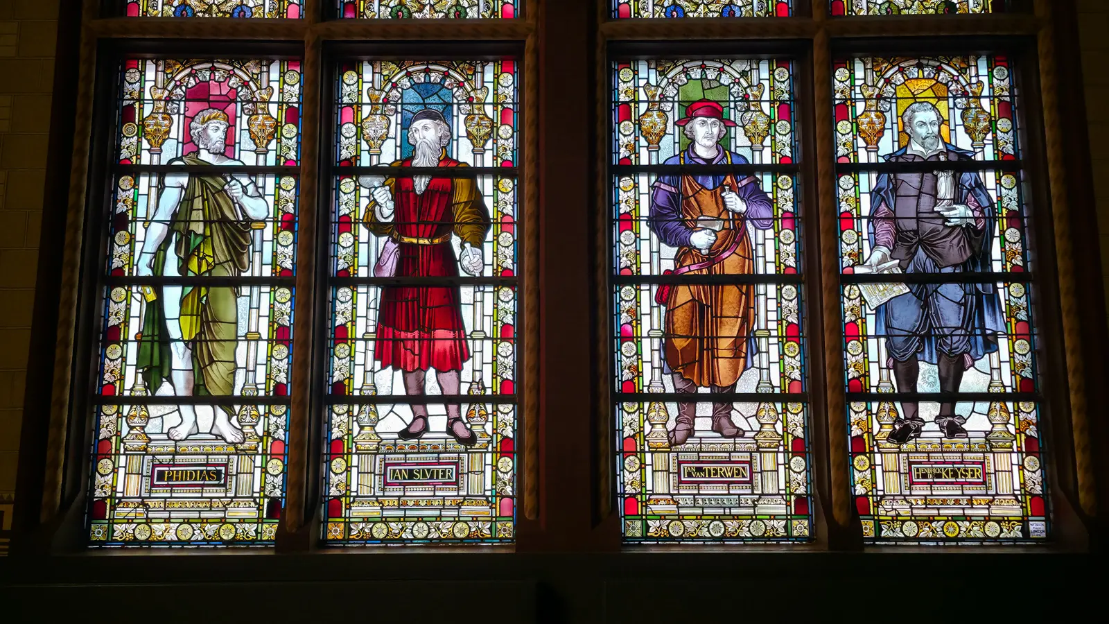 Stained glass windows at the Rijksmuseum, four panels each showing a standing figure with a name lettered on the pedestal below