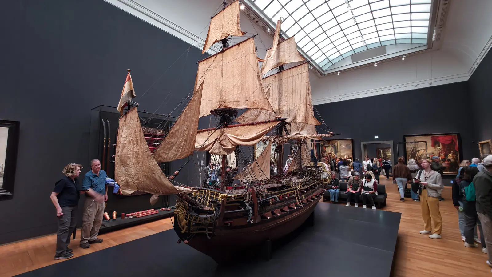 A large rigged model ship with sails set, on a low plinth in a skylit gallery at the Rijksmuseum, with paintings on the walls beyond