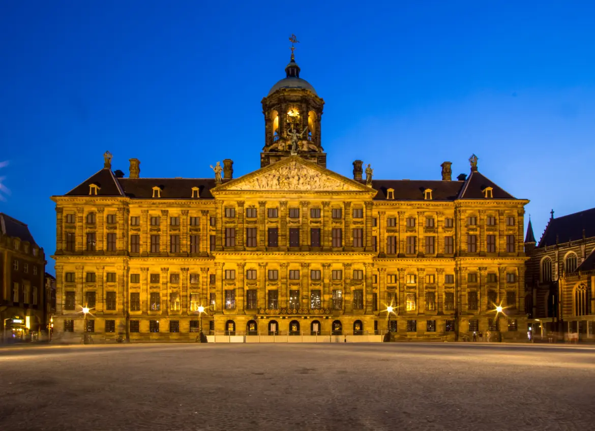 The Royal Palace of Amsterdam floodlit at dusk on Dam Square, with its cupola, pediment and weathervane against a blue sky