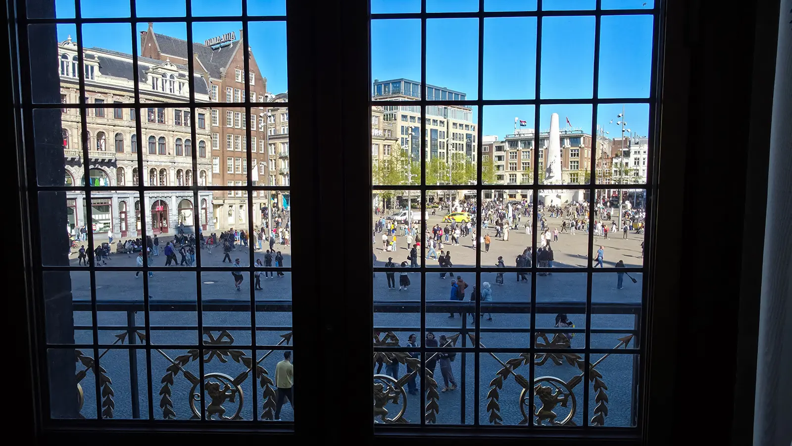 Dam Square in Amsterdam seen through a leaded window of the Royal Palace, with gilded ironwork across the lower panes and the National Monument beyond