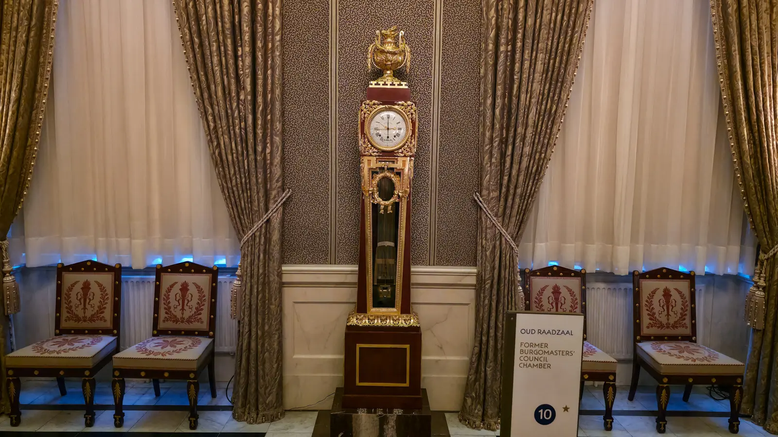 A tall gilt and mahogany longcase clock in the Oud Raadzaal of the Royal Palace Amsterdam, flanked by chairs beneath draped curtains