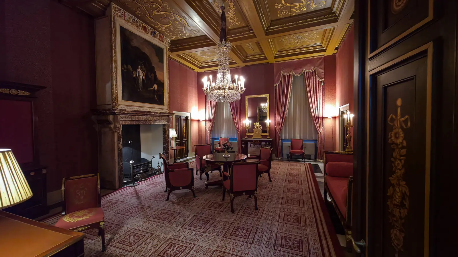 A red room in the Royal Palace Amsterdam with a gilded coffered ceiling, chandelier, marble chimneypiece and Empire seating around a round table