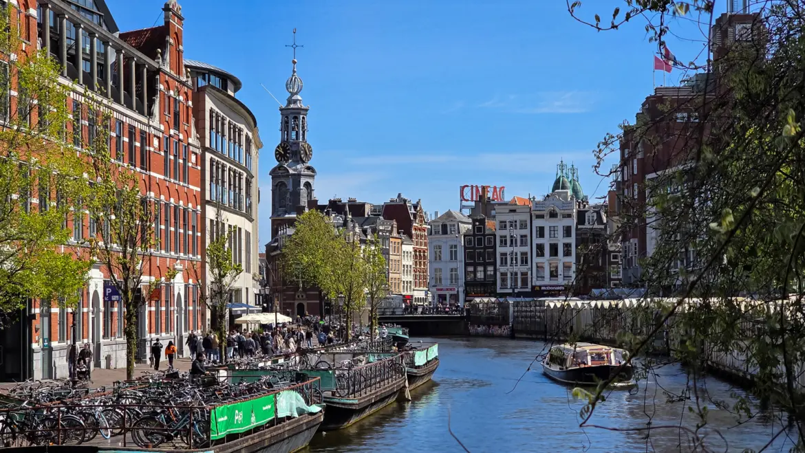 The Singel canal in Amsterdam looking towards the Munttoren, with moored barges, bicycles, flower-market awnings and a tour boat
