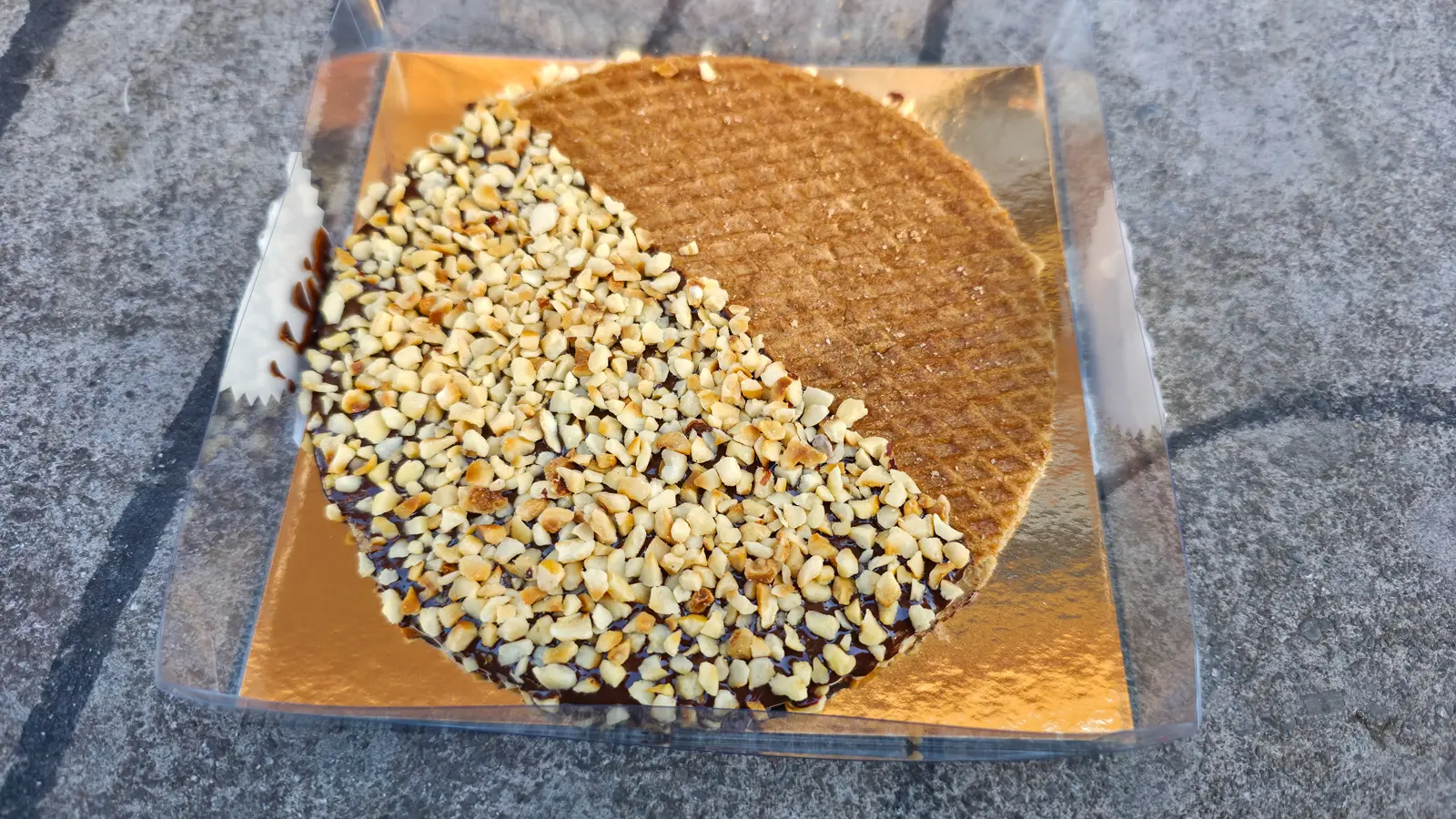 A stroopwafel half-covered in dark chocolate and chopped hazelnuts on a gold board on a grey stone surface in Amsterdam