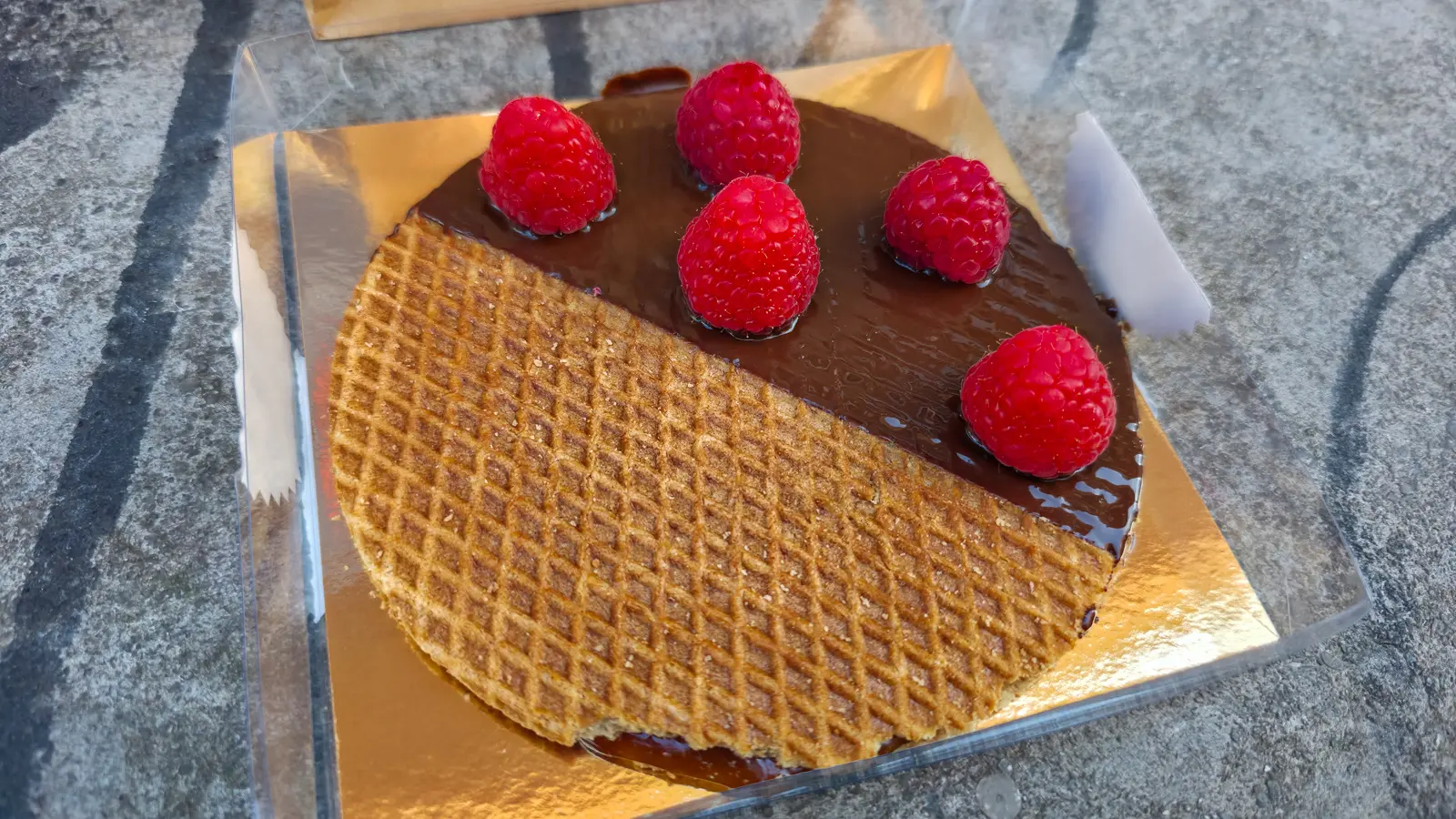 A stroopwafel topped with dark chocolate and five fresh raspberries on a gold board on a grey stone surface in Amsterdam