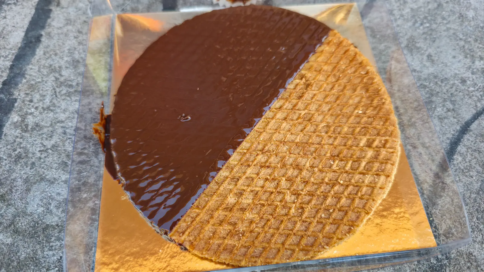 A stroopwafel half-coated in dark chocolate on a gold board, photographed on a grey stone surface in Amsterdam