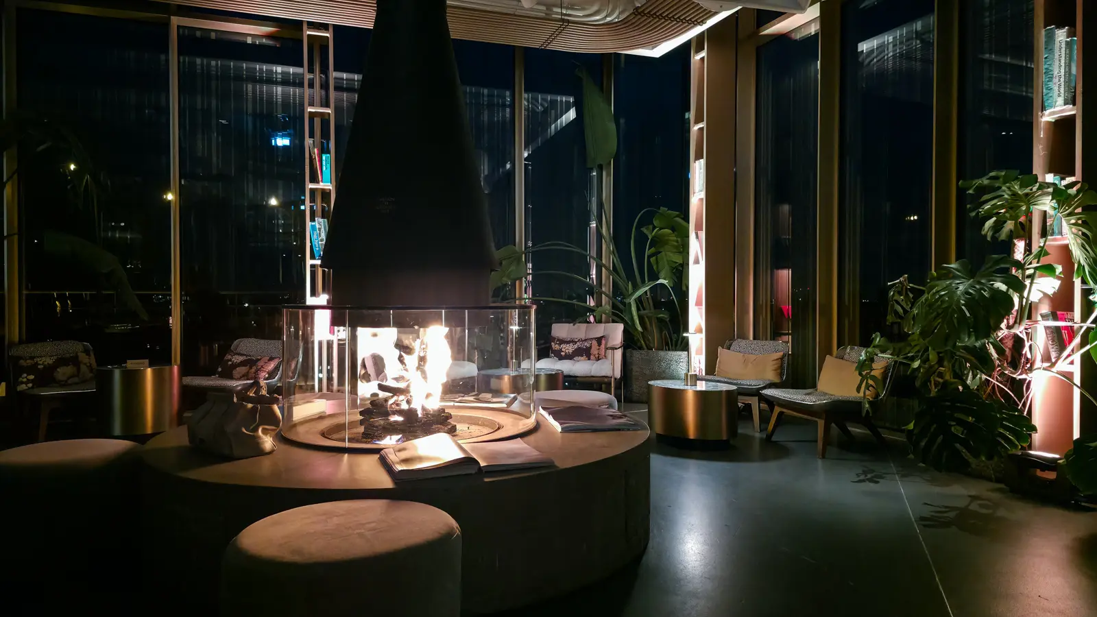 The conical fireplace lit at night in the W Lounge at the W Amsterdam, with lamps, plants and the dark city beyond the glass