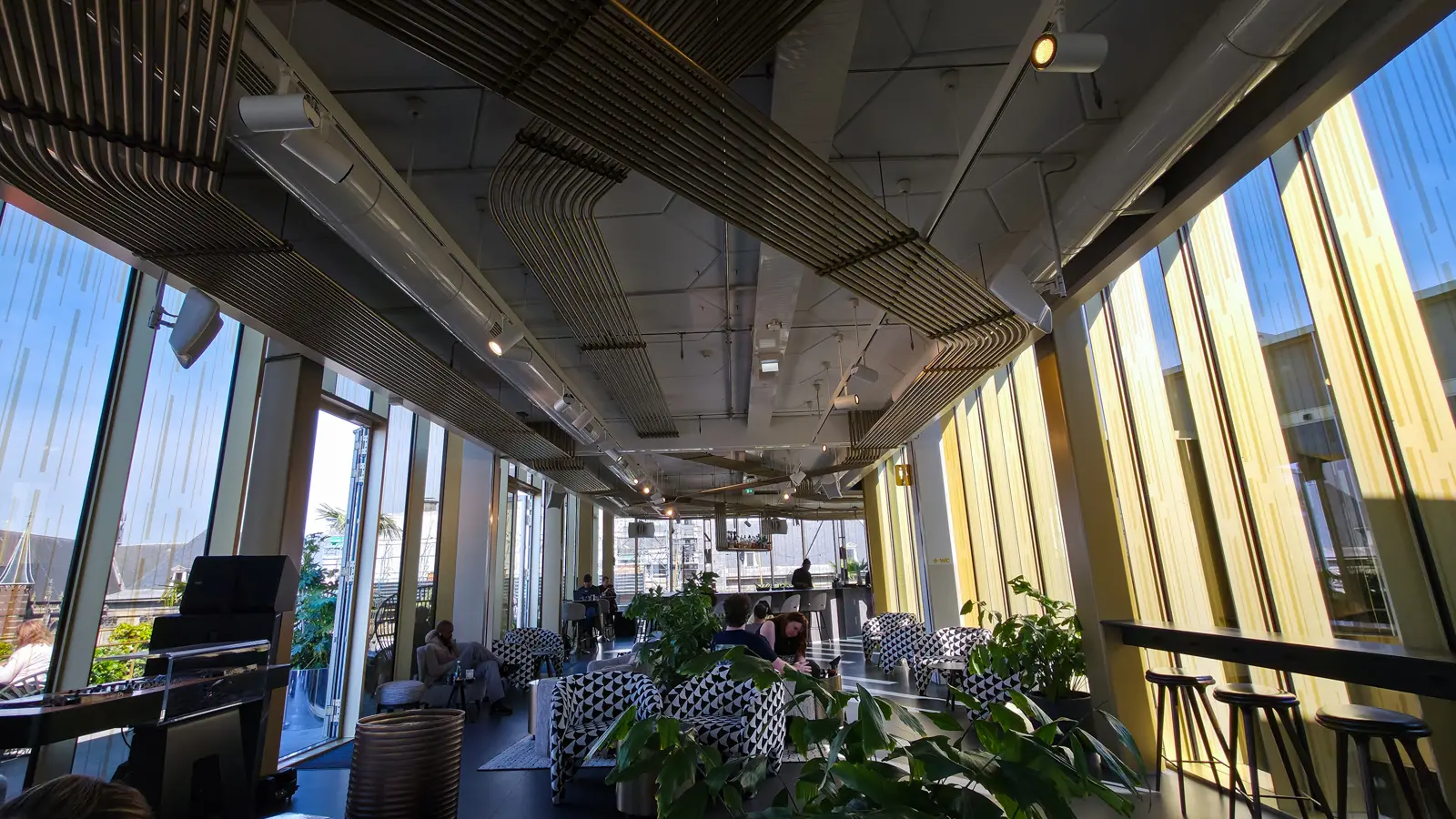 The long glass-walled interior of the W Lounge at the W Amsterdam, with bar stools, plants and exposed ducting overhead