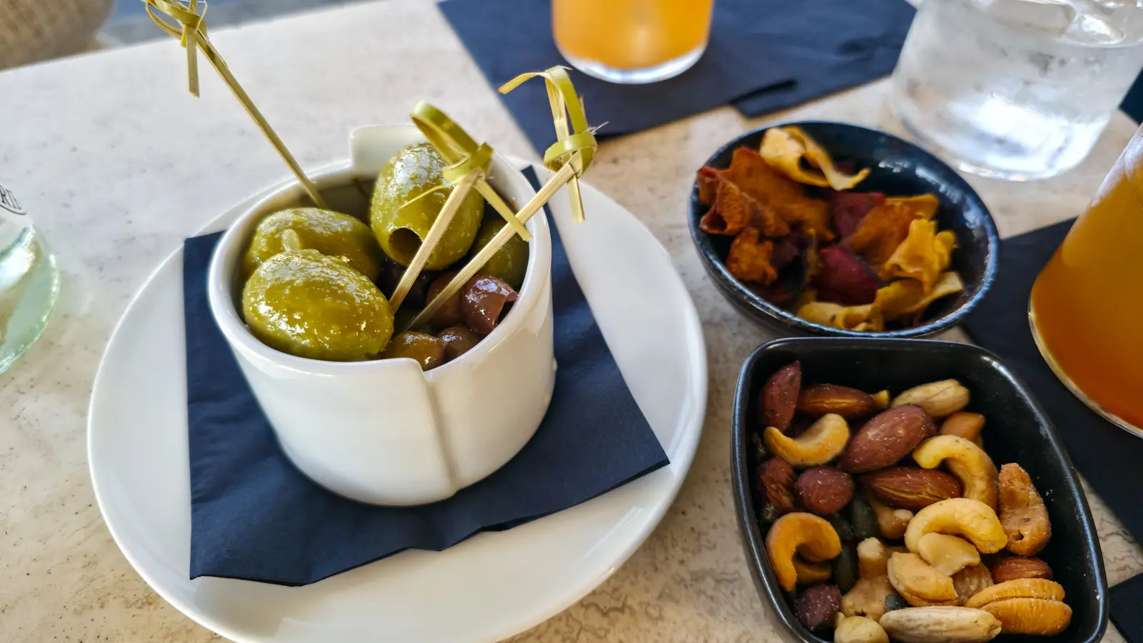 Marinated green and dark olives in a divided white pot with wooden picks, beside bowls of nuts and vegetable crisps at the W Lounge