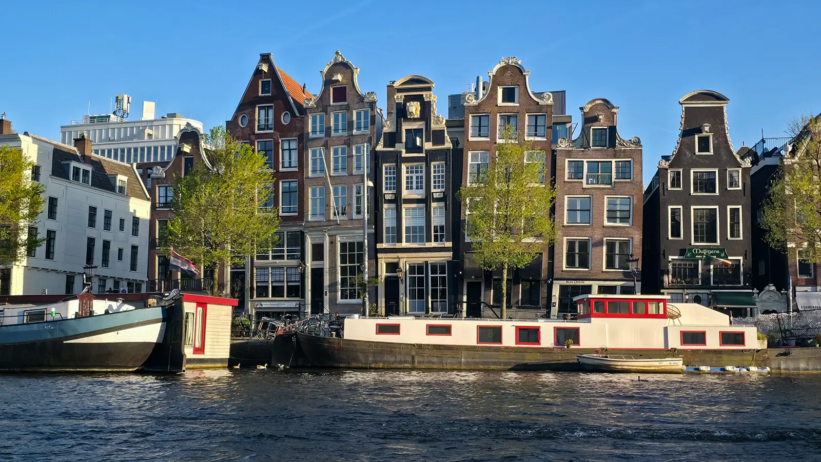 A row of gabled canal houses on the Amstel in Amsterdam, leaning at different angles in low golden light, with houseboats moored in front