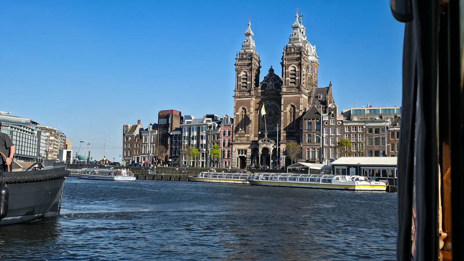 The twin towers of the Basilica of Saint Nicholas above the Amsterdam waterfront, with canal tour boats moored below