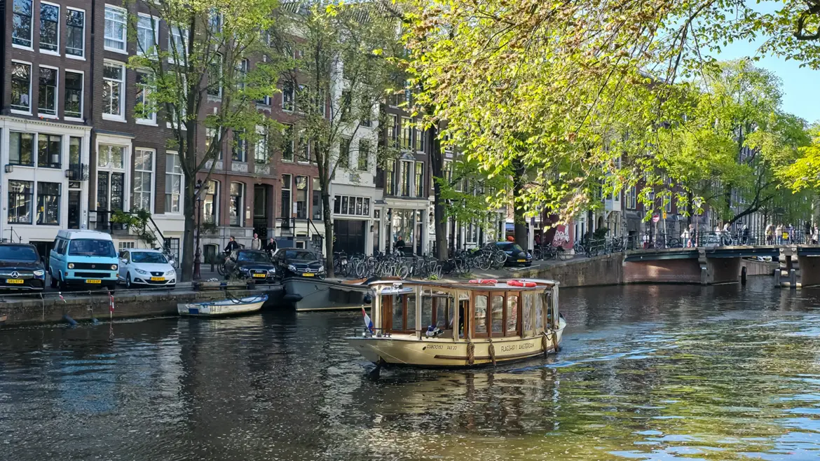 Small covered saloon boat on an Amsterdam canal, cream hull and varnished timber window frames, with lifebuoys on the canopy and a Dutch flag at the bow