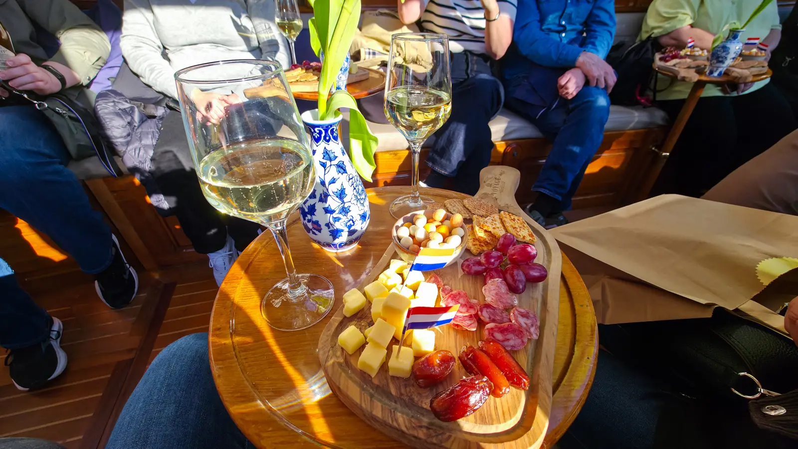 Dutch snack board on a canal boat with cheese cubes, dry sausage, grapes, dates and stroopwafels, a Delft vase and two glasses of white wine