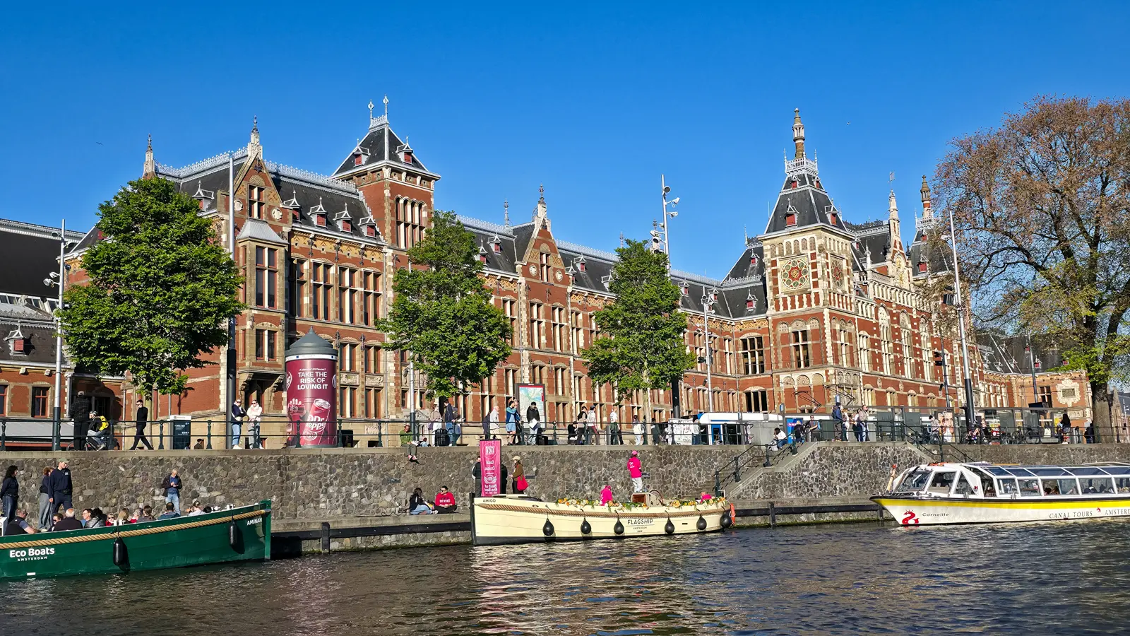 Amsterdam Centraal station seen from the water, its full red-brick facade visible at once, with tour boats moored along the quay in front