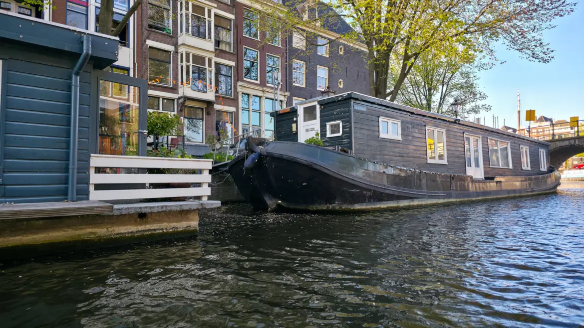 An old black barge converted into a houseboat on an Amsterdam canal, beside a grey wooden houseboat with a white fence