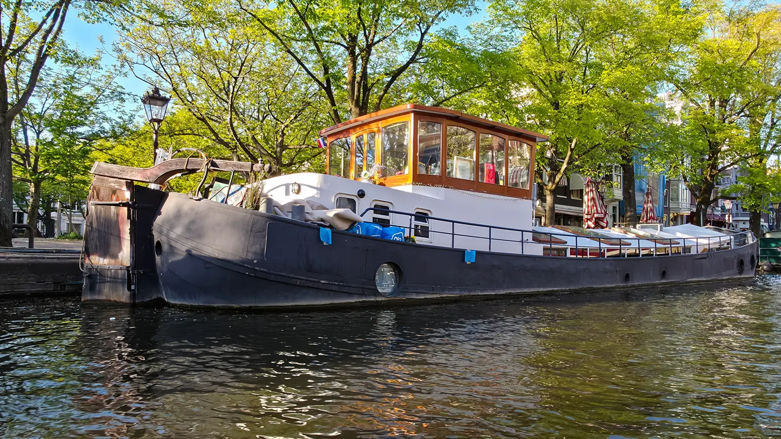 Black-hulled houseboat with a varnished timber wheelhouse glowing in the evening sun, moored under trees on an Amsterdam canal