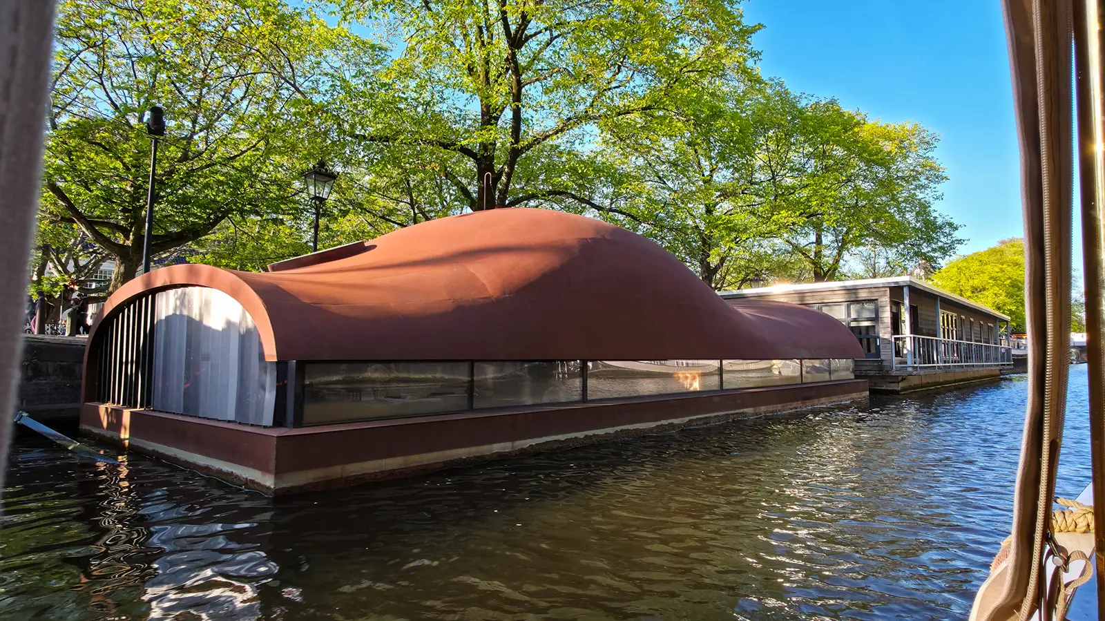 A modern houseboat with a curved rust-red metal roof and a band of windows, moored under trees on an Amsterdam canal