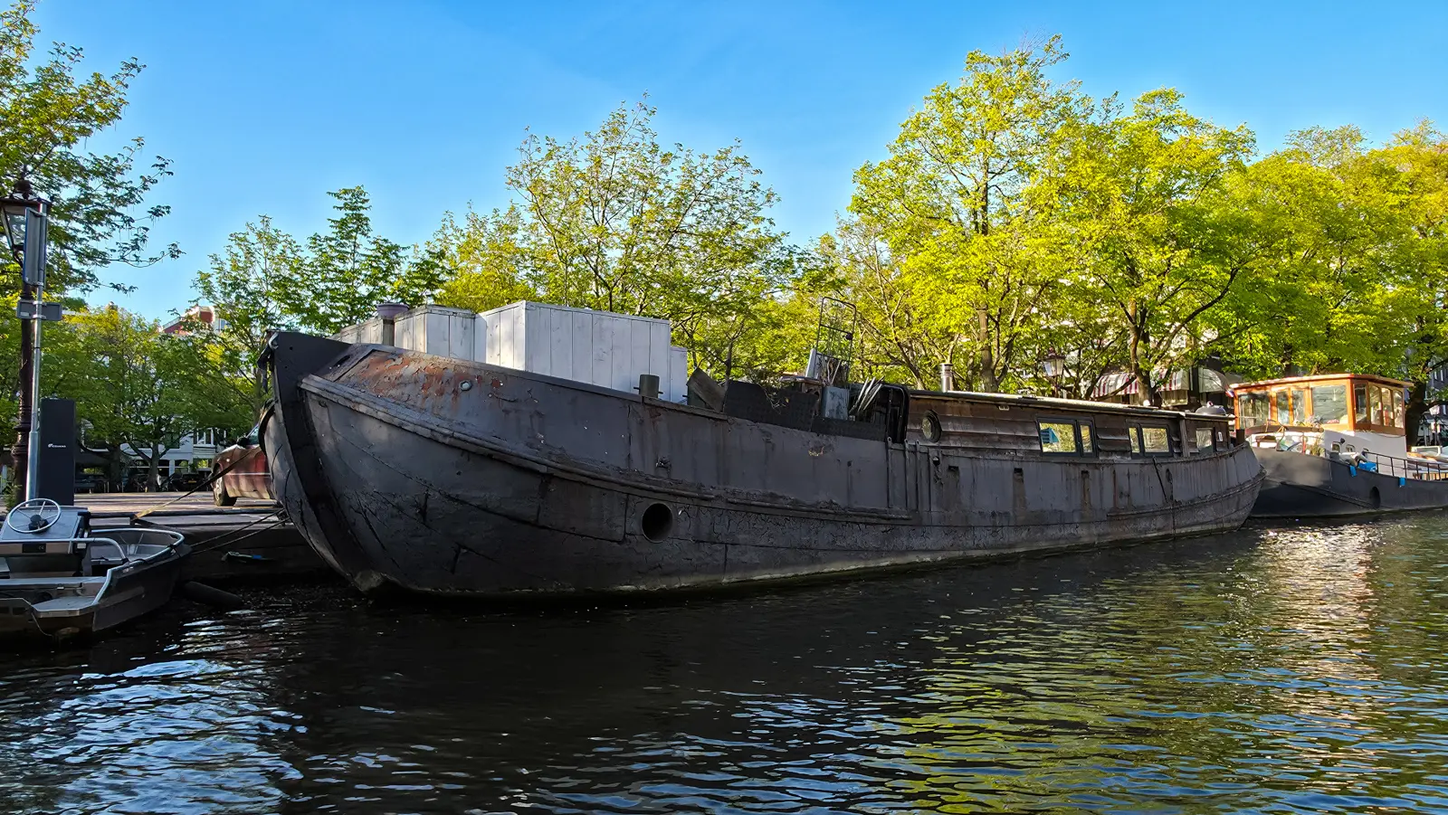 A weathered steel barge with a porthole and white timber boxes on deck, moored on an Amsterdam canal in evening light